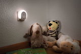 Weißer LED-Nachtlicht-Stecker an Wand mit Kuscheltieren und Kinderbüchern im Zimmer.