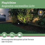 Schwarzer Gartenstrahler mit warmweißem Licht beleuchtet grüne Pflanzen neben Terrasse mit Sofa