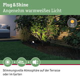 Schwarzer Gartenstrahler mit warmweißem Licht beleuchtet grüne Pflanzen neben Terrasse mit Sofa