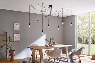 Salle à manger moderne avec table en bois clair, chaises blanches et suspension noire avec ampoules.