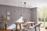 Salle à manger moderne avec table en bois clair, chaises blanches et suspension noire avec ampoules.