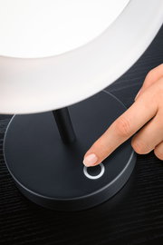 Finger berührt den runden Touch-Schalter einer schwarzen Tischlampe mit weißem Lampenschirm auf dunklem Holz.