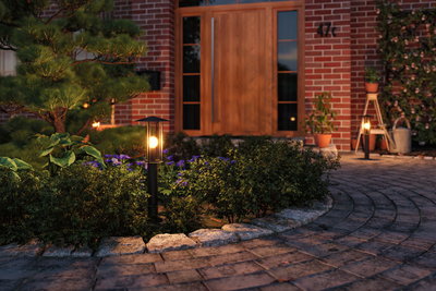 Black outdoor lamp with warm white light in garden by front door with stone path and plants.