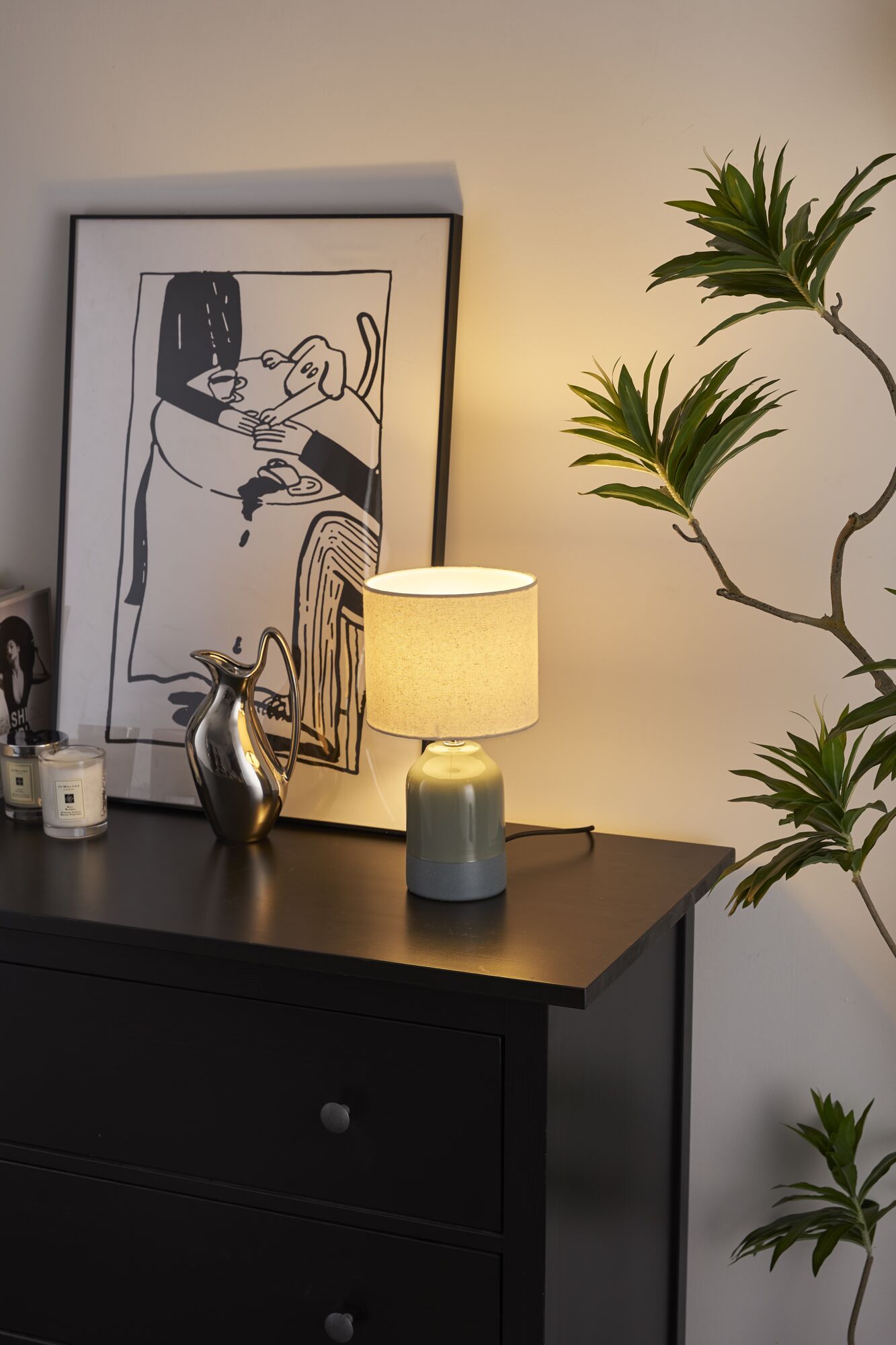 Grijze keramische tafellamp met lichte stoffen kap op zwarte ladekast naast moderne kunst en groene plant