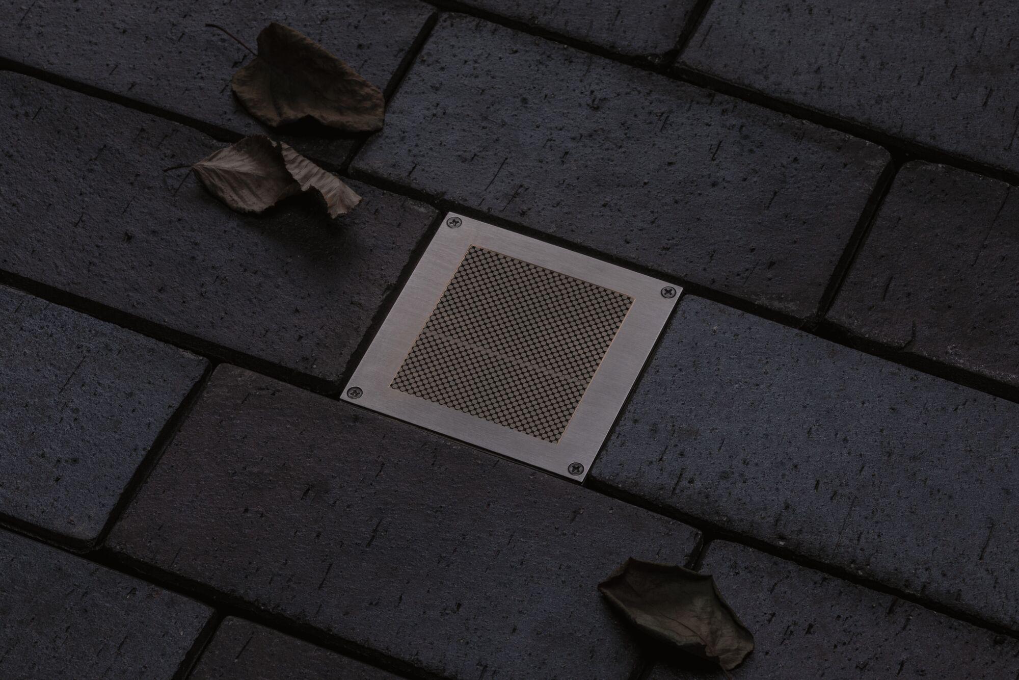 Square stainless steel floor light with mesh insert on dark paving stones with autumn leaves