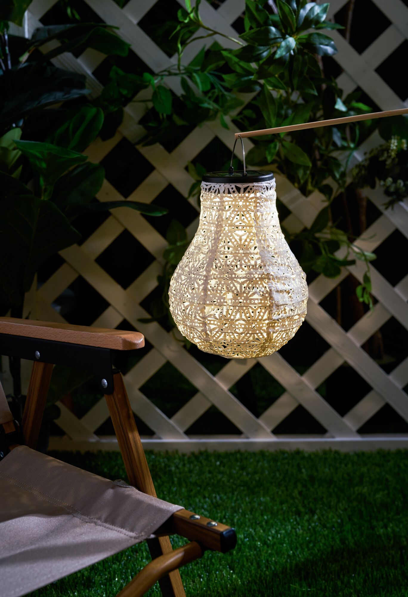 Verlichte witte stoffen lantaarn met patroon hangend in tuin naast houten stoel en groen gras.