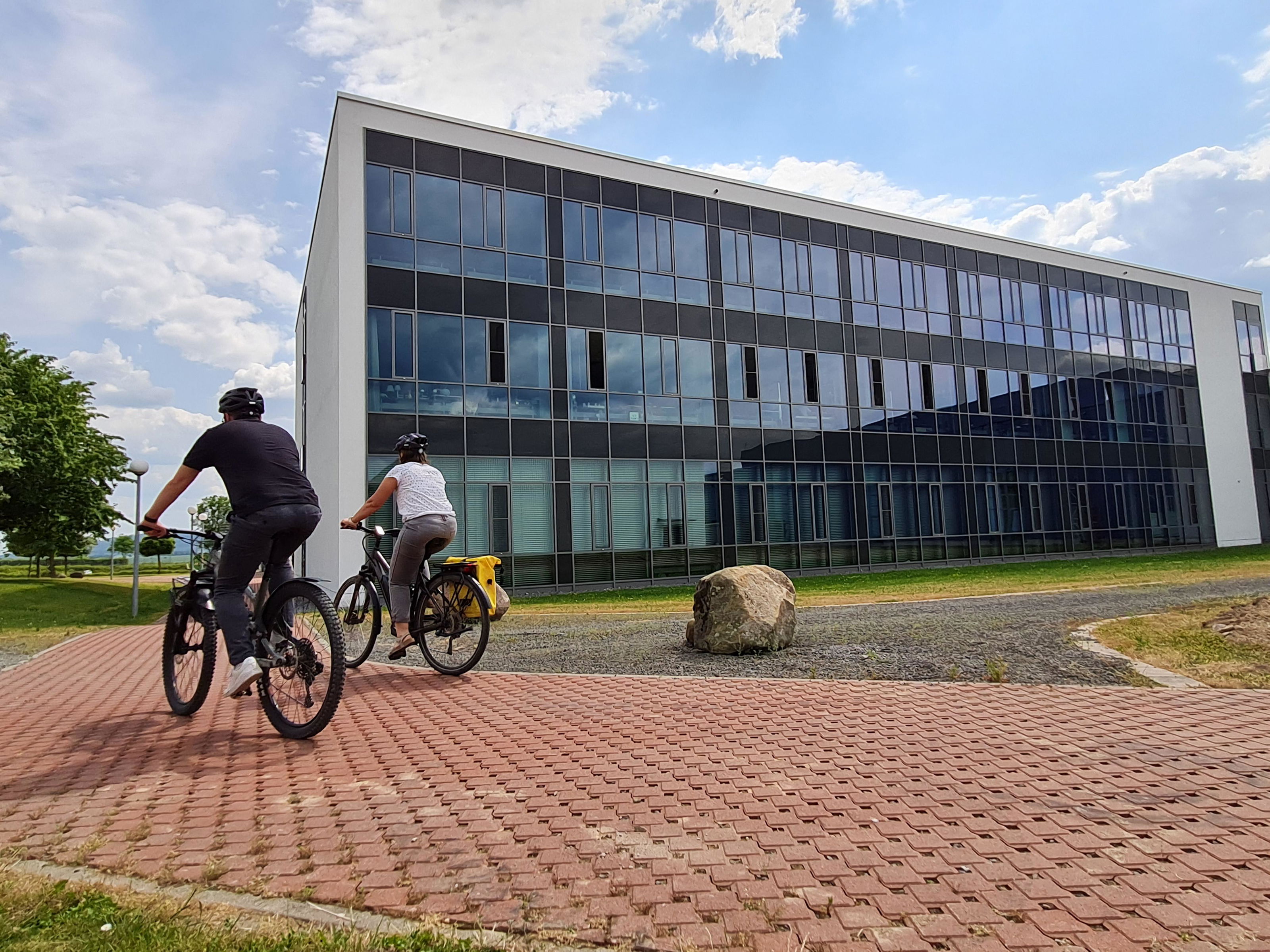 Two cyclists wearing helmets ride on red pavement in front of a modern office building with large glass windows.