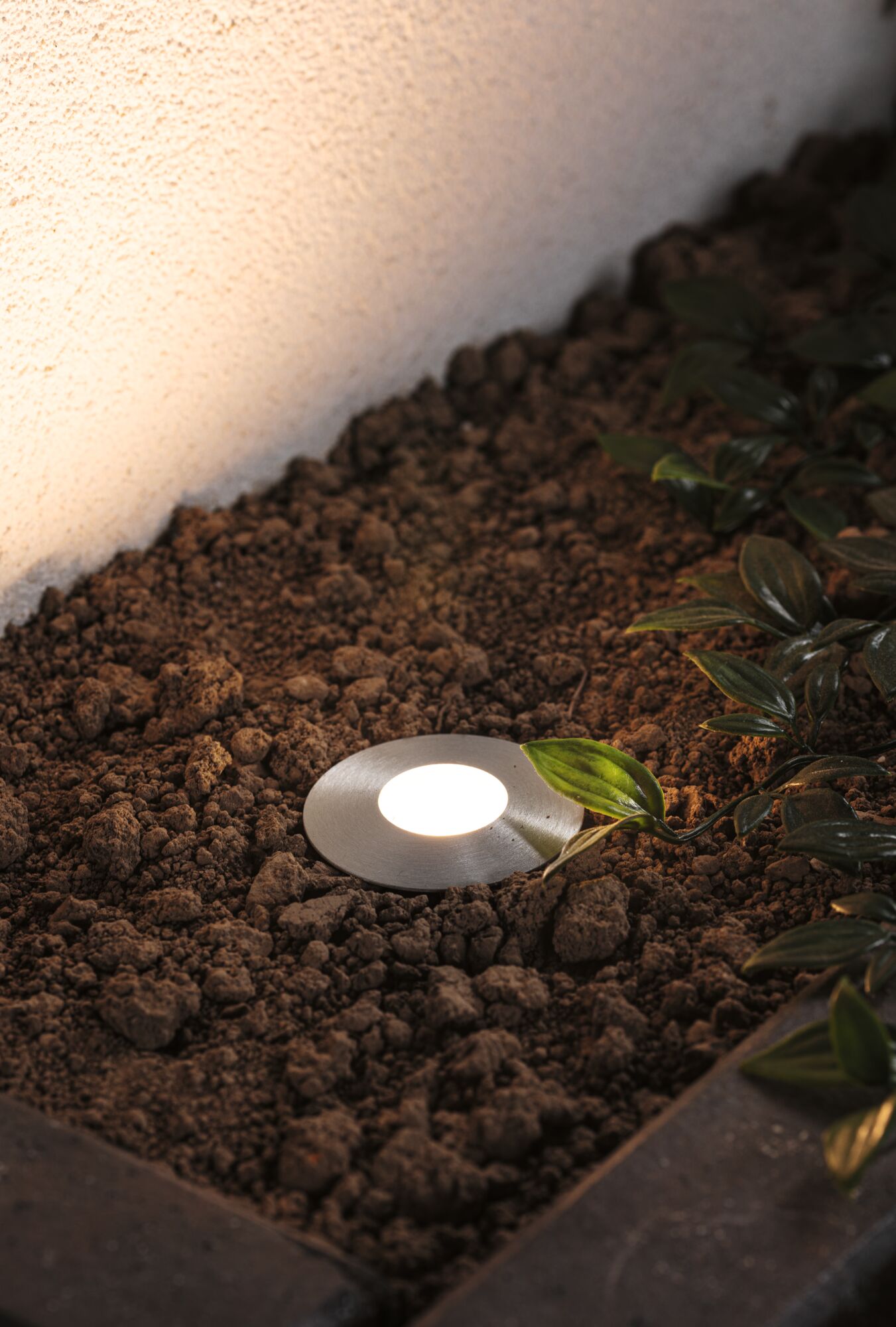 Rundes Edelstahl-Einbauleuchte mit warmweißem LED-Licht im Gartenboden neben grünen Blättern.