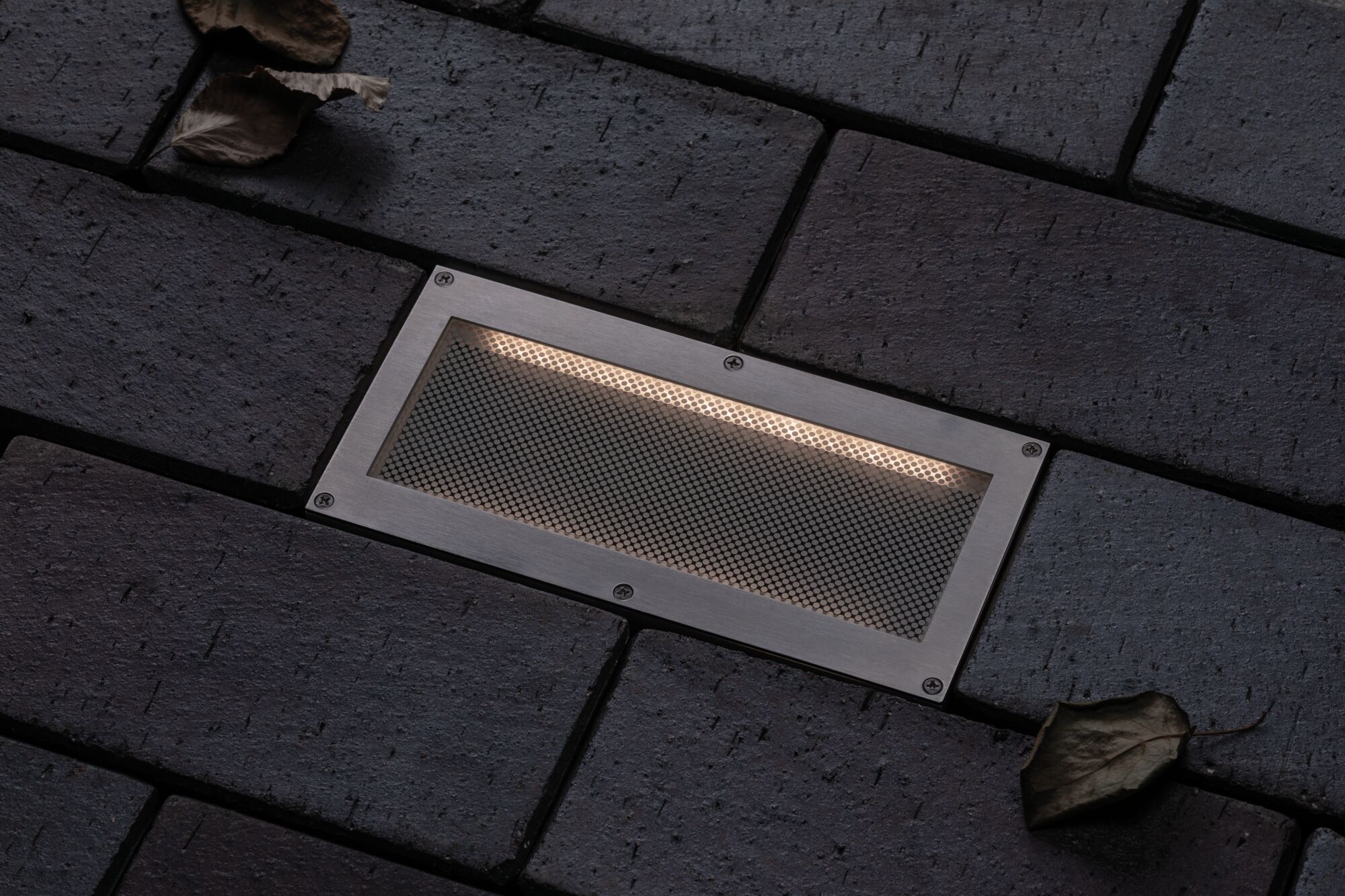 Eingelassene LED-Bodenleuchte aus gebürstetem Edelstahl mit warmweißem Licht auf dunklem Pflaster.