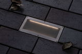 Recessed LED floor light in brushed stainless steel with warm white light on dark paving stones.
