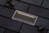 Recessed LED floor light in brushed stainless steel with warm white light on dark paving stones.