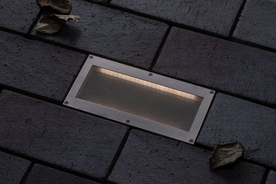 Recessed LED floor light in brushed stainless steel with warm white light on dark paving stones.