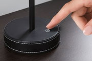 Schwarzer runder Lampenfuß mit Lederbezug und weißer Naht, Finger berührt kleinen Schalter.