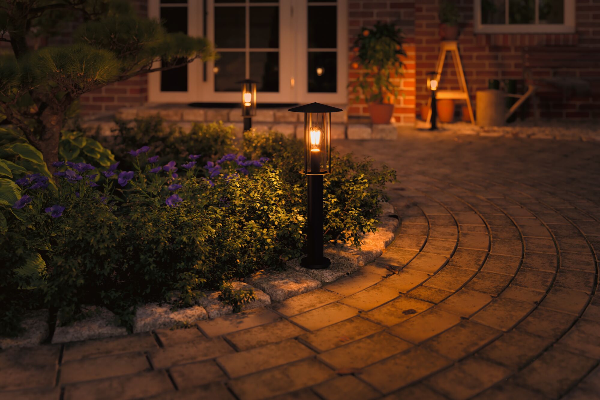 Schwarze Außenleuchte aus Metall mit warmweißem Licht am Gartenweg neben Blumenbeet bei Abenddämmerung
