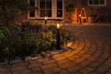 Black metal outdoor lamp with warm white light along garden path near flower bed at dusk