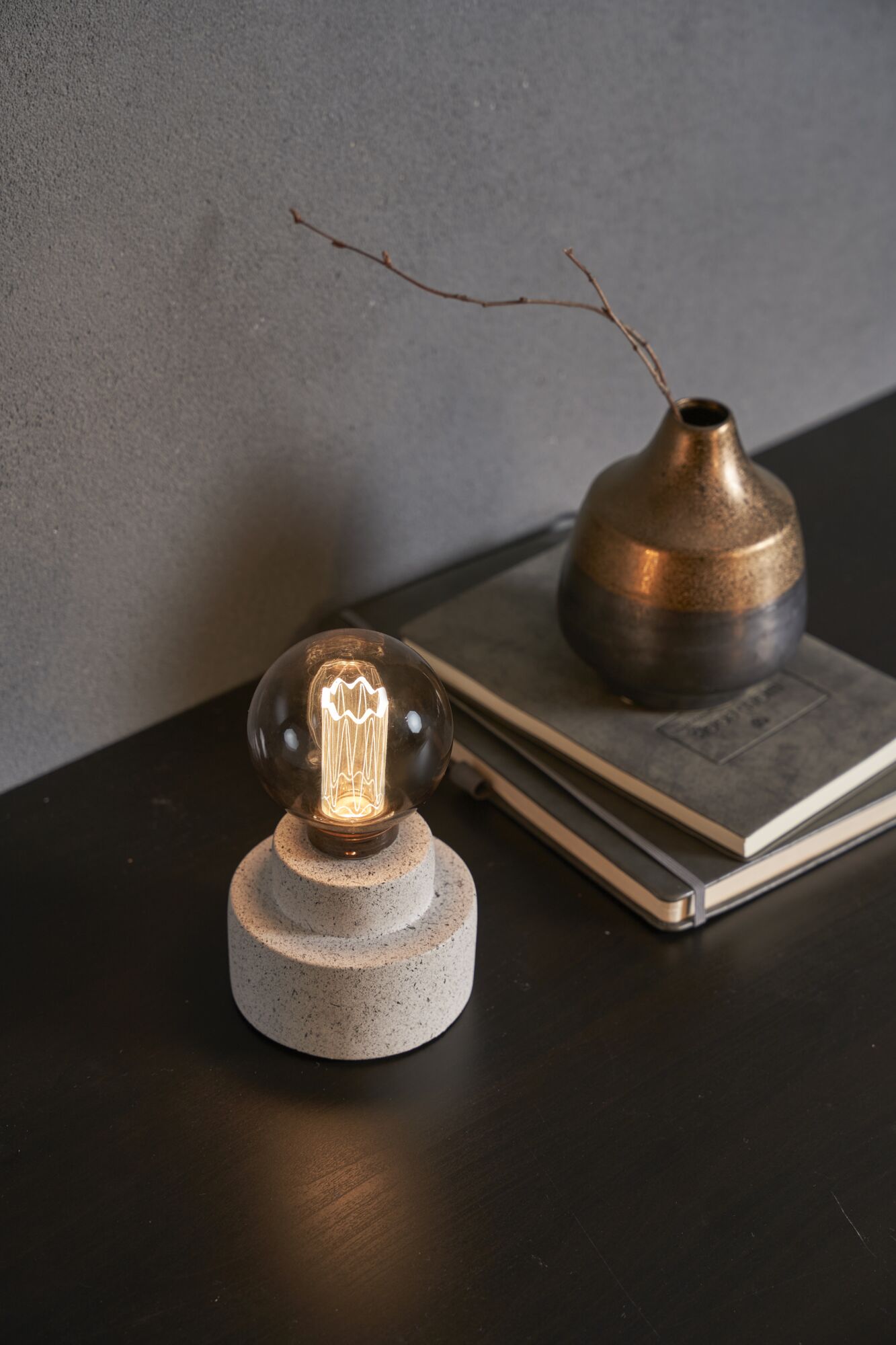 Modern table lamp with round bulb and grey stone base on dark wood table next to decorative vase.