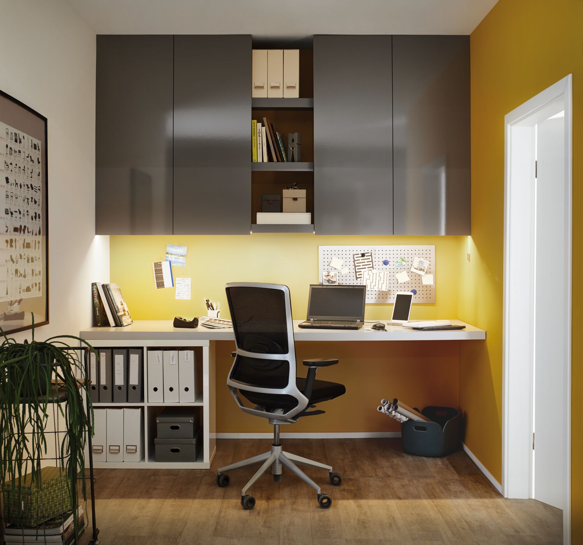 Moderne thuiswerkplek met grijze hangkasten, gele muur, ergonomische bureaustoel en LED-werkverlichting