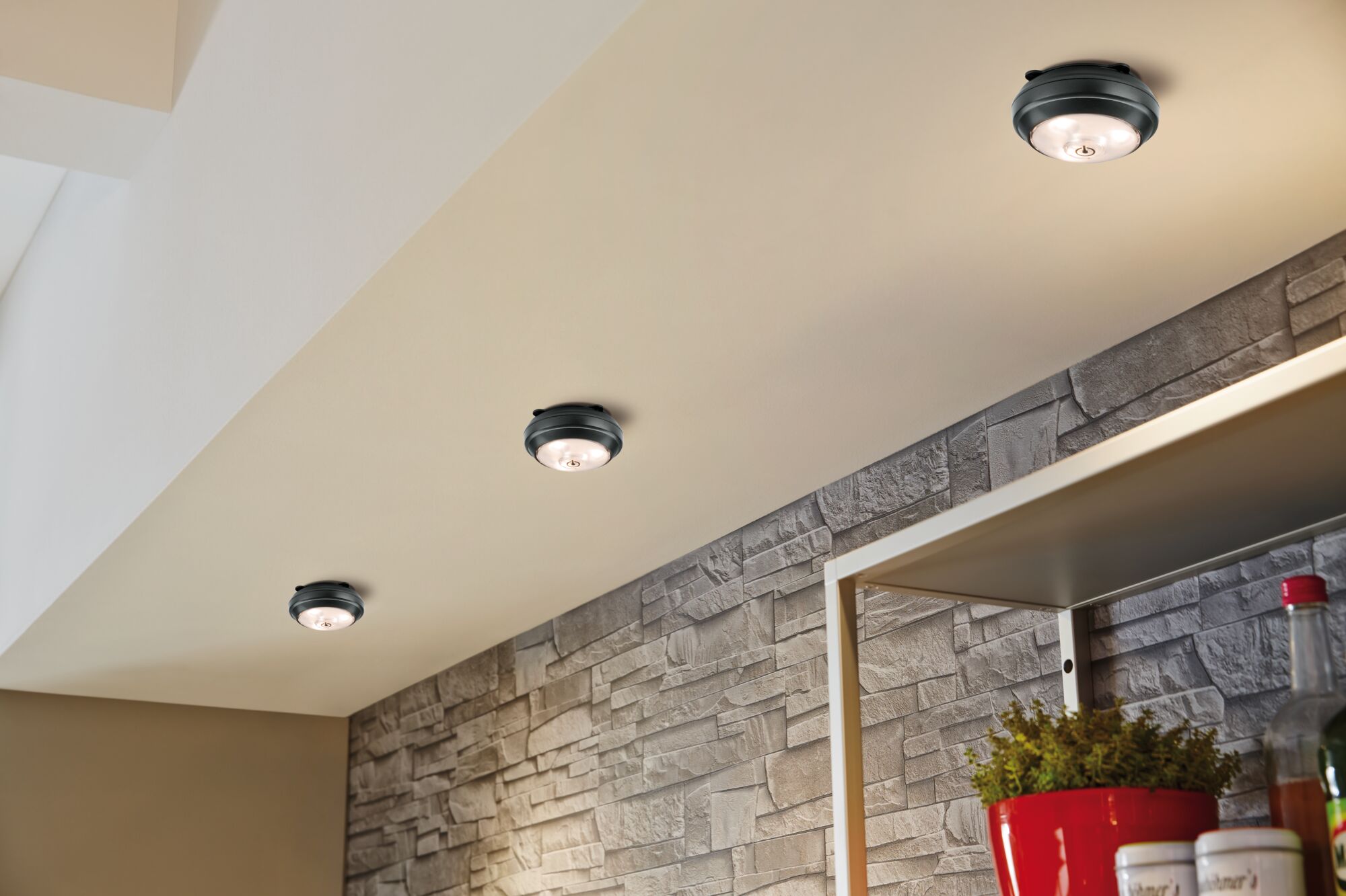 Three black LED ceiling lights with warm white light on beige ceiling above stone wall and shelf.
