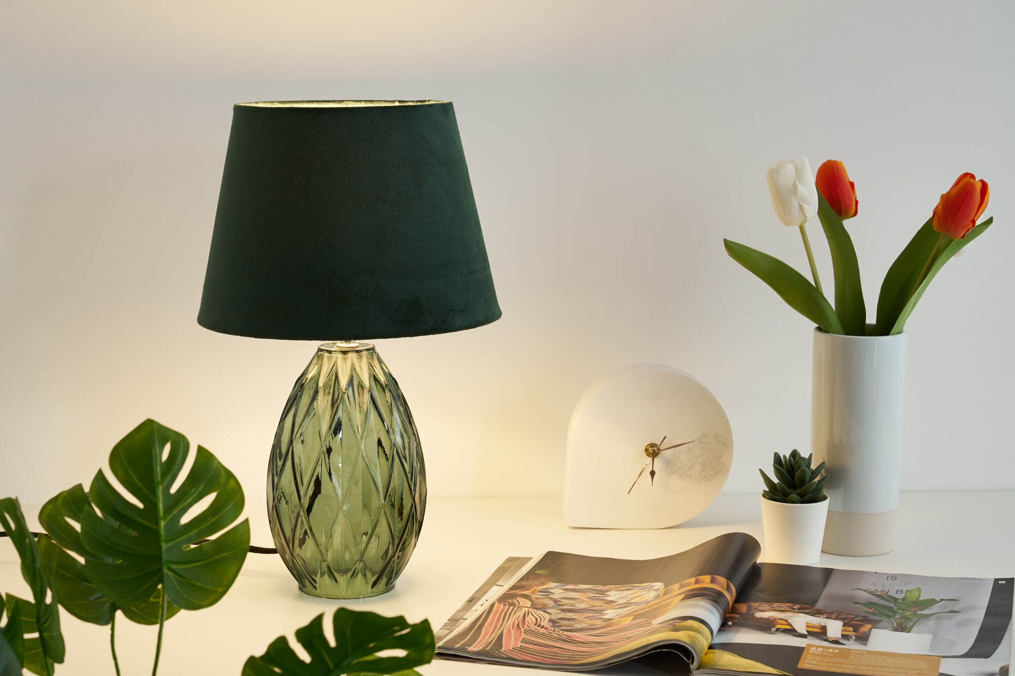 Lampe de table en verre vert avec abat-jour en tissu vert foncé à côté d'une horloge blanche et de tulipes sur une table blanche