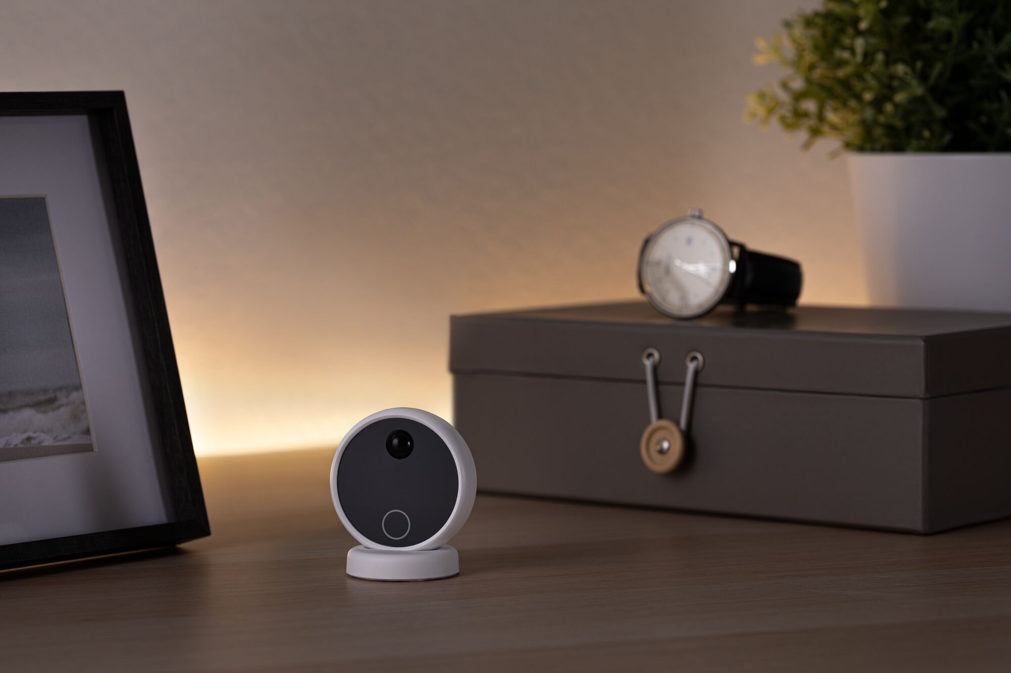 Small white security camera with black front on wooden table near watch and gray box