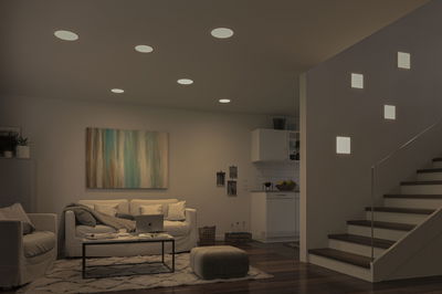 Modern living room with white LED recessed lights and square wall lamps by staircase, wooden flooring