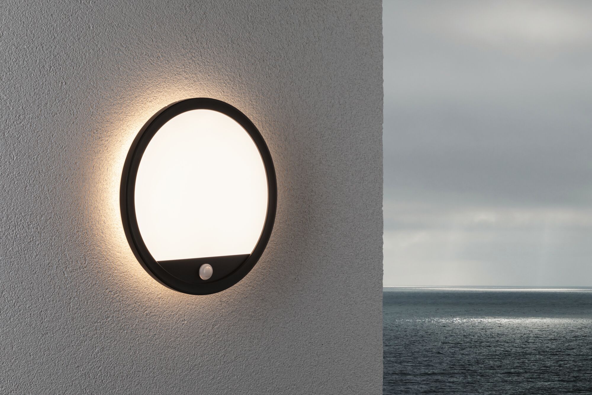 Round LED wall light with black frame and motion sensor on grey wall overlooking the sea