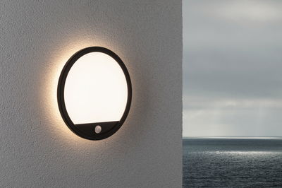 Round LED wall light with black frame and motion sensor on grey wall overlooking the sea