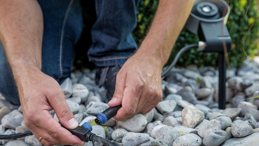 Mann verbindet schwarze Outdoor-Lichtkabel auf grauem Kies, Gartenbeleuchtung Installation.