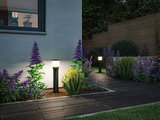 Modern black outdoor lights with warm white glow illuminate garden path with purple flowers and stone wall.
