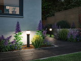 Moderne zwarte buitenlampen met warm wit licht verlichten tuinpad met paarse bloemen en stenen muur.