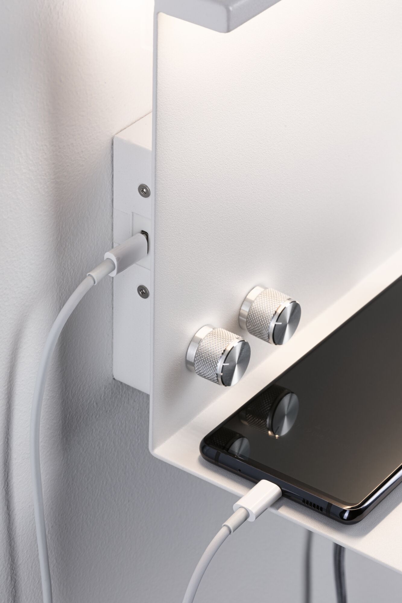 Weißes Wandregal mit zwei silbernen Drehknöpfen und Smartphone, das per USB-Kabel geladen wird