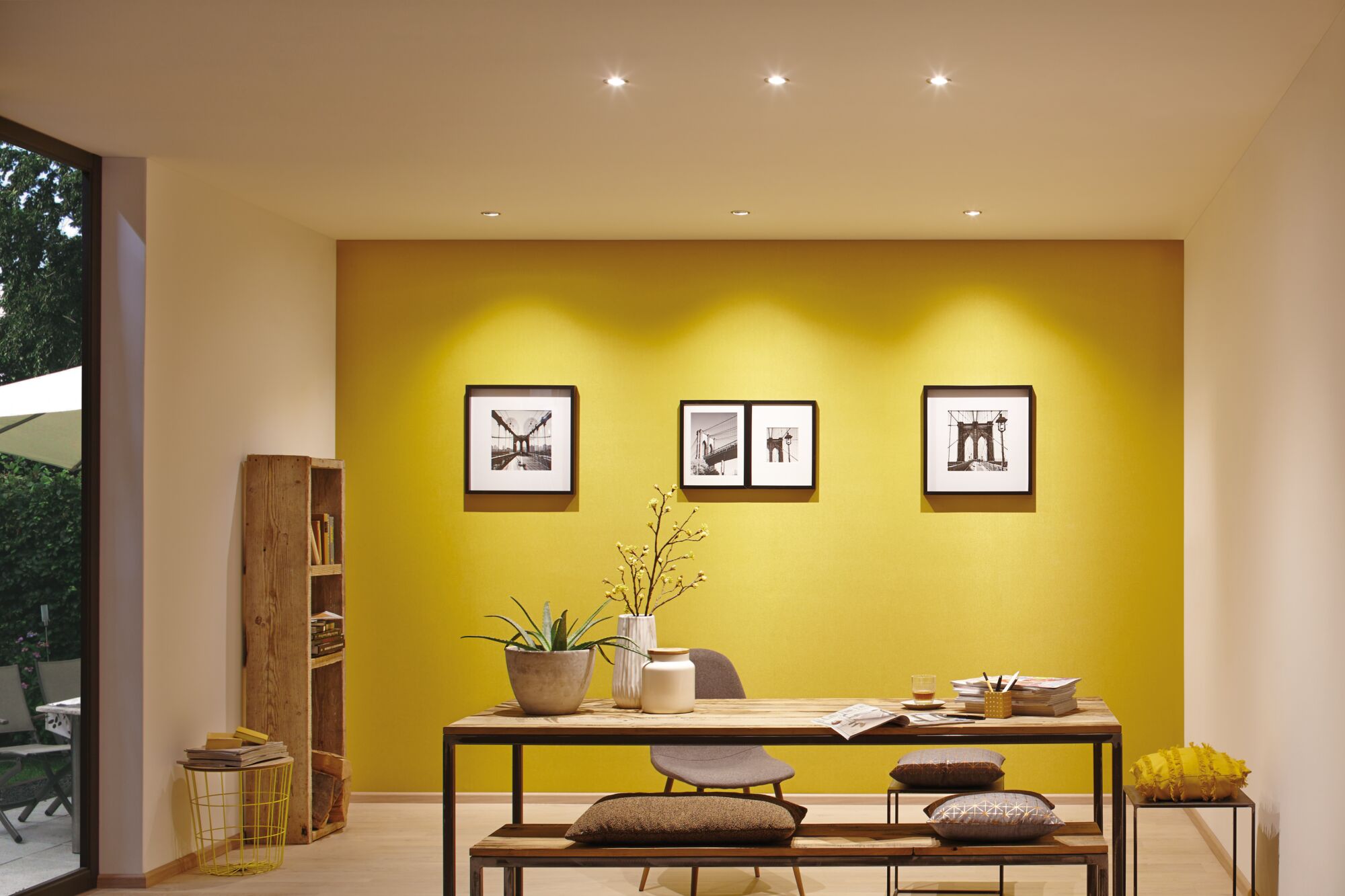 Moderne eetkamer met gele muur, houten bank, tafel, decoratieve kussens en LED-spotverlichting.
