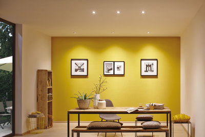 Moderne eetkamer met gele muur, houten bank, tafel, decoratieve kussens en LED-spotverlichting.