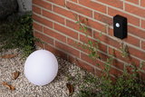 White plastic globe light on gravel by red brick wall with black motion sensor for outdoor lighting.