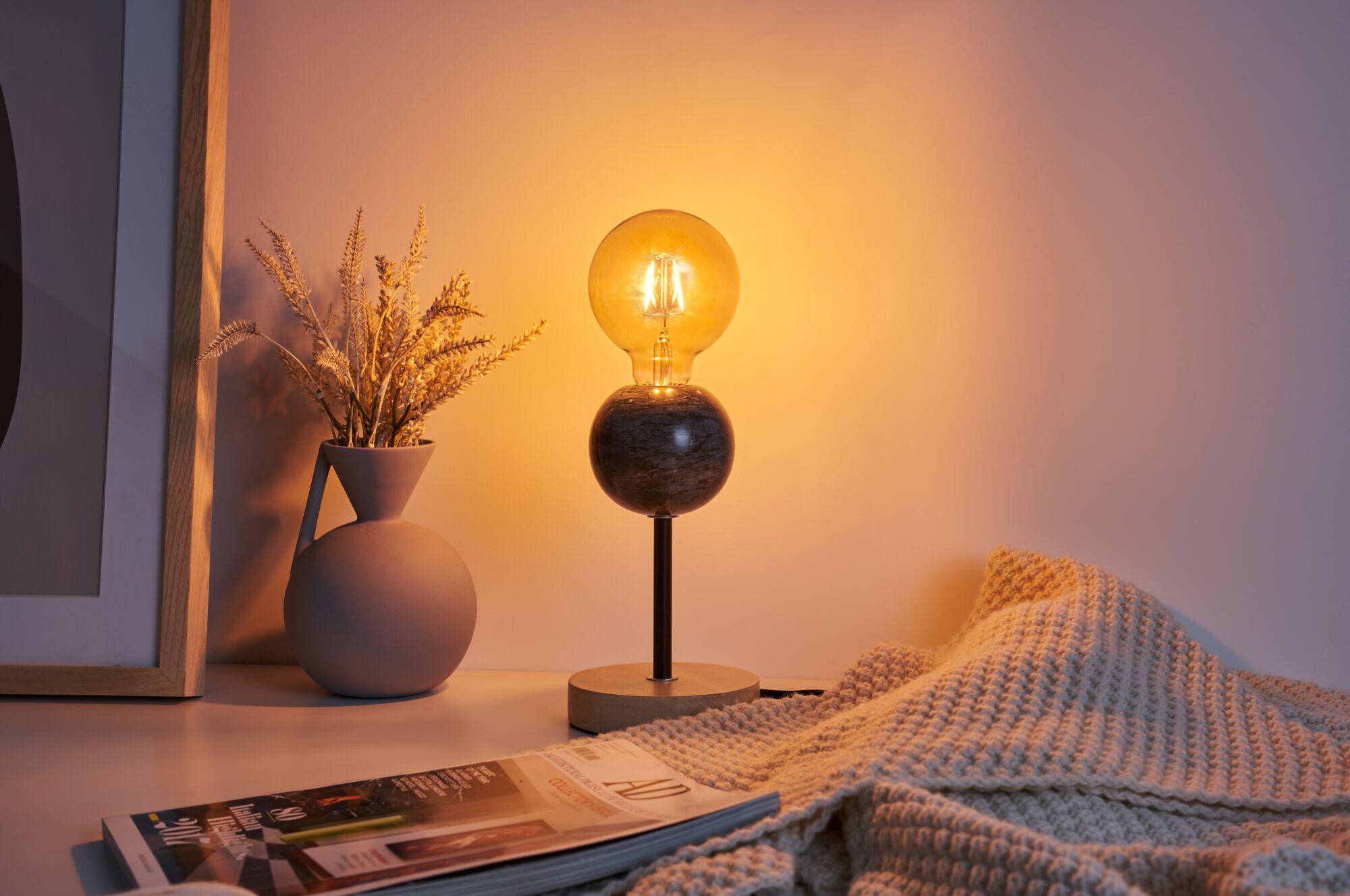 Modern table lamp with wooden base and black rod, warm light beside grey vase and blanket