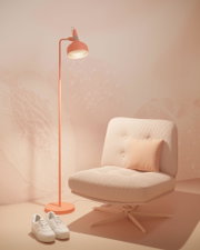 Pomarańczowa lampa stojąca z metalu obok kremowego fotela z poduszką i białych trampek