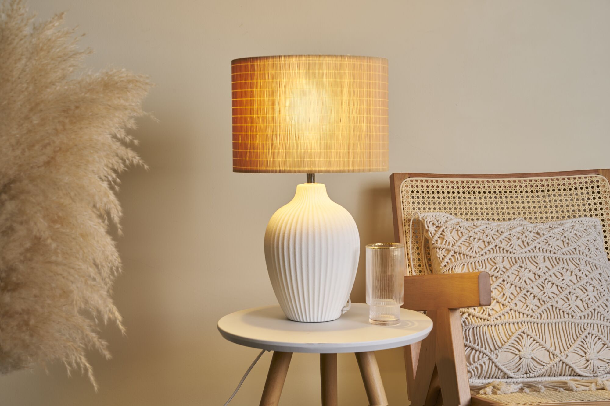 Lampe de table en céramique blanche à design côtelé avec lumière chaude sur table ronde en bois près d'un fauteuil.