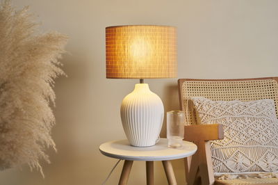 Lampe de table en céramique blanche à design côtelé avec lumière chaude sur table ronde en bois près d'un fauteuil.