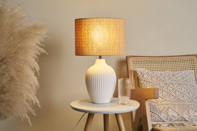 Lampe de table en céramique blanche à design côtelé avec lumière chaude sur table ronde en bois près d'un fauteuil.