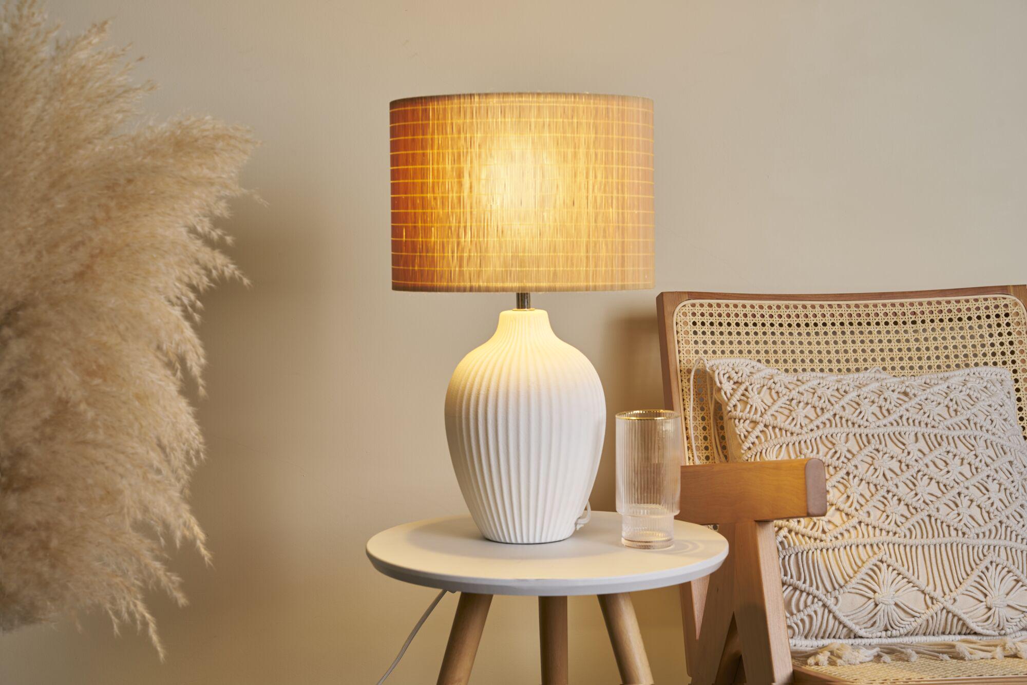 Lampe de table en céramique blanche avec abat-jour en tissu marron sur table ronde en bois à côté d'une chaise en rotin