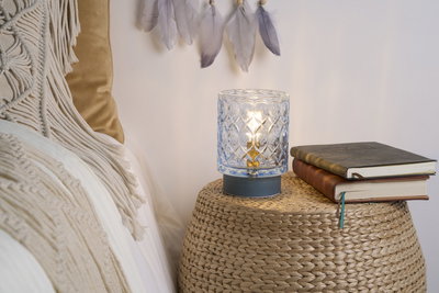 Glazen tafellamp met patroon en warm licht op gevlochten bijzettafel naast boeken en beddengoed