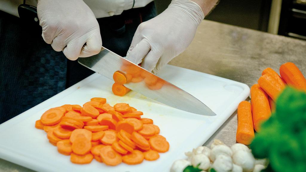 Person schneidet frische orange Karotten auf weißem Schneidebrett in Profi-Küchenumgebung.