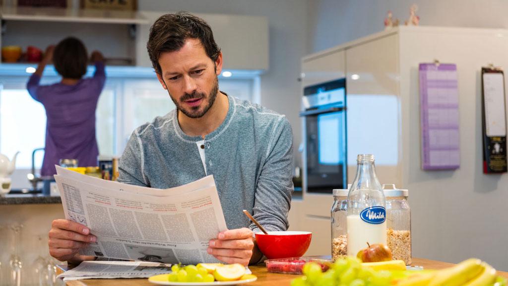 Mann liest Zeitung am Küchentisch mit Milchflasche und Obst, Alltagsszene in moderner Küche.