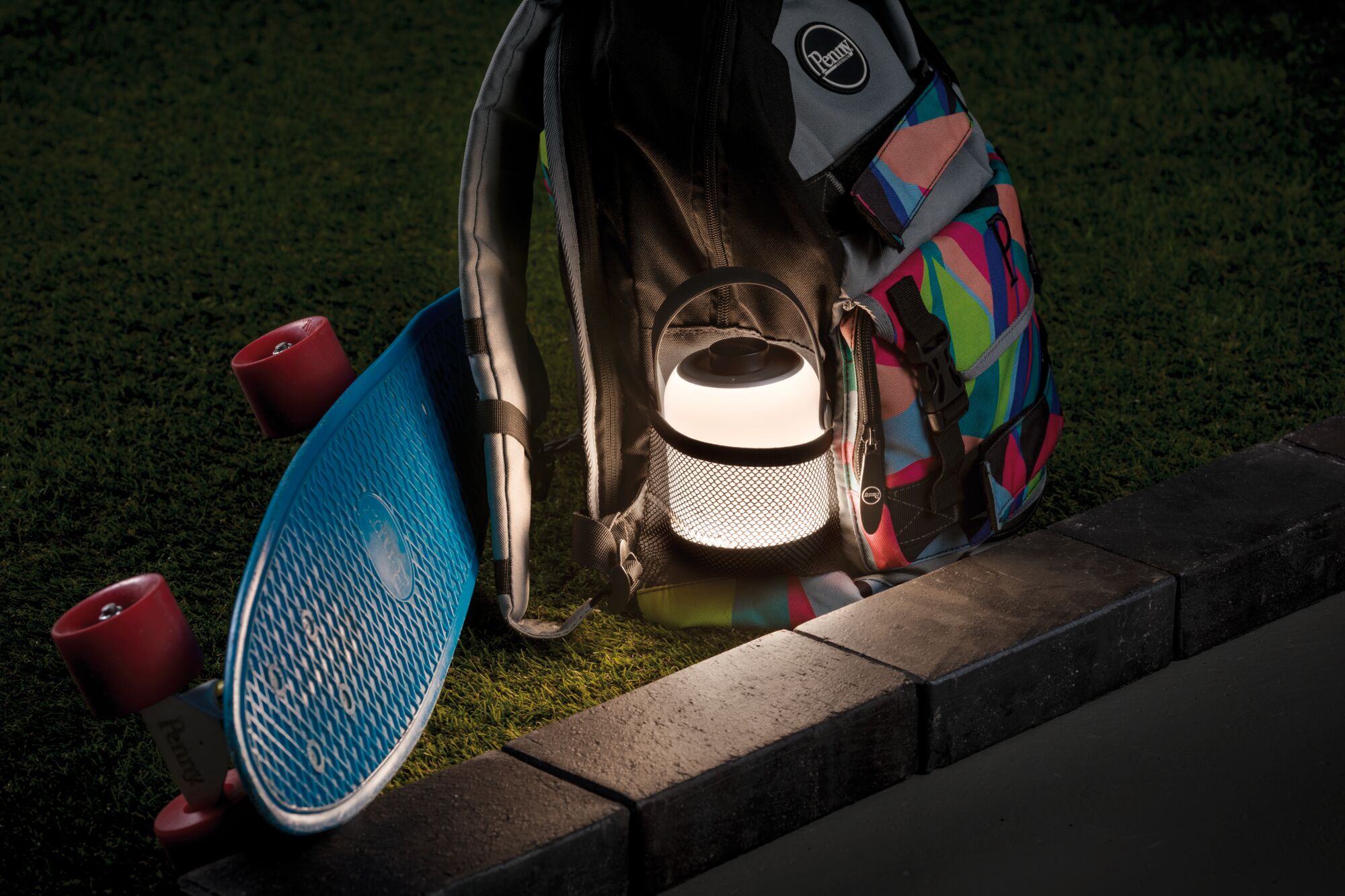 Leuchtende tragbare LED-Laterne neben buntem Rucksack und blauem Skateboard auf Gras bei Nacht