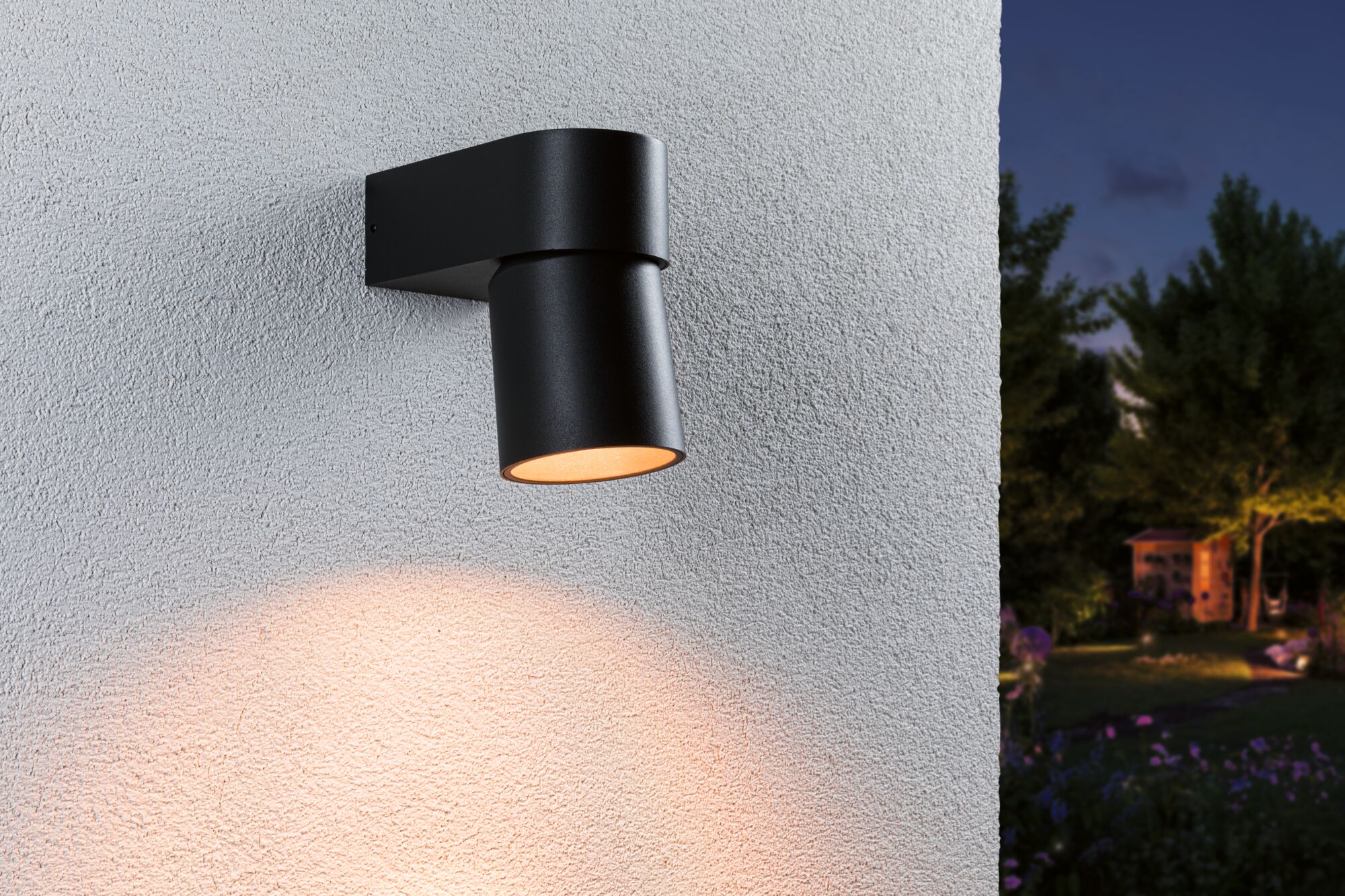 Black metal outdoor wall light with warm white illumination on white wall in garden at night