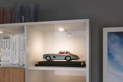Zilveren modelauto op zwarte standaard verlicht in witte kast met designboeken en glazen vazen.
