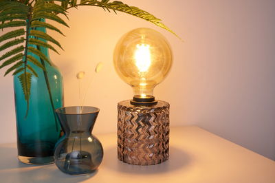 Tafellamp met gestructureerde glazen voet en grote lamp naast blauwe glazen vazen en varenblad.