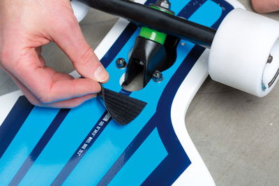 Main fixant une bande auto-agrippante noire sur un skateboard bleu et blanc avec roues blanches et amortisseur vert