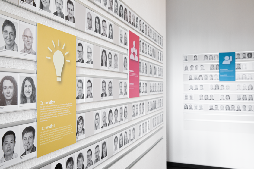 Wall displaying numerous employee portraits with colorful info panels about company values like innovation and cooperation