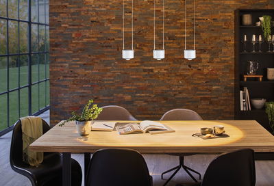 Drie witte LED-hanglampen boven lichte houten tafel met boeken en kopje in eetkamer met bakstenen muur